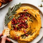 Bowl of creamy pumpkin hummus garnished with herbs and served with fresh vegetables.